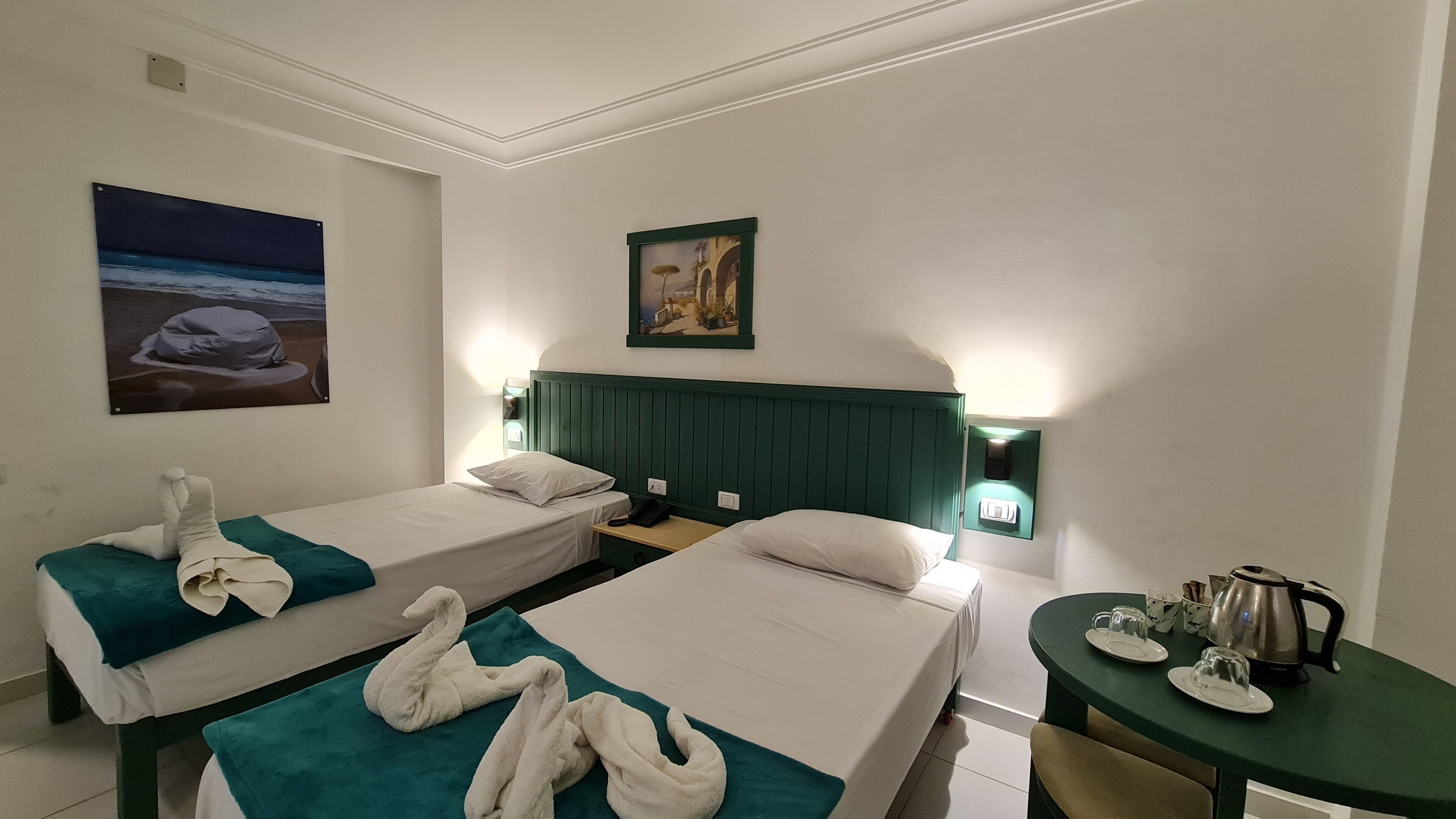 A hotel room with two single beds, a green headboard, a painting of a beach scene, and a small table with a kettle and cups.