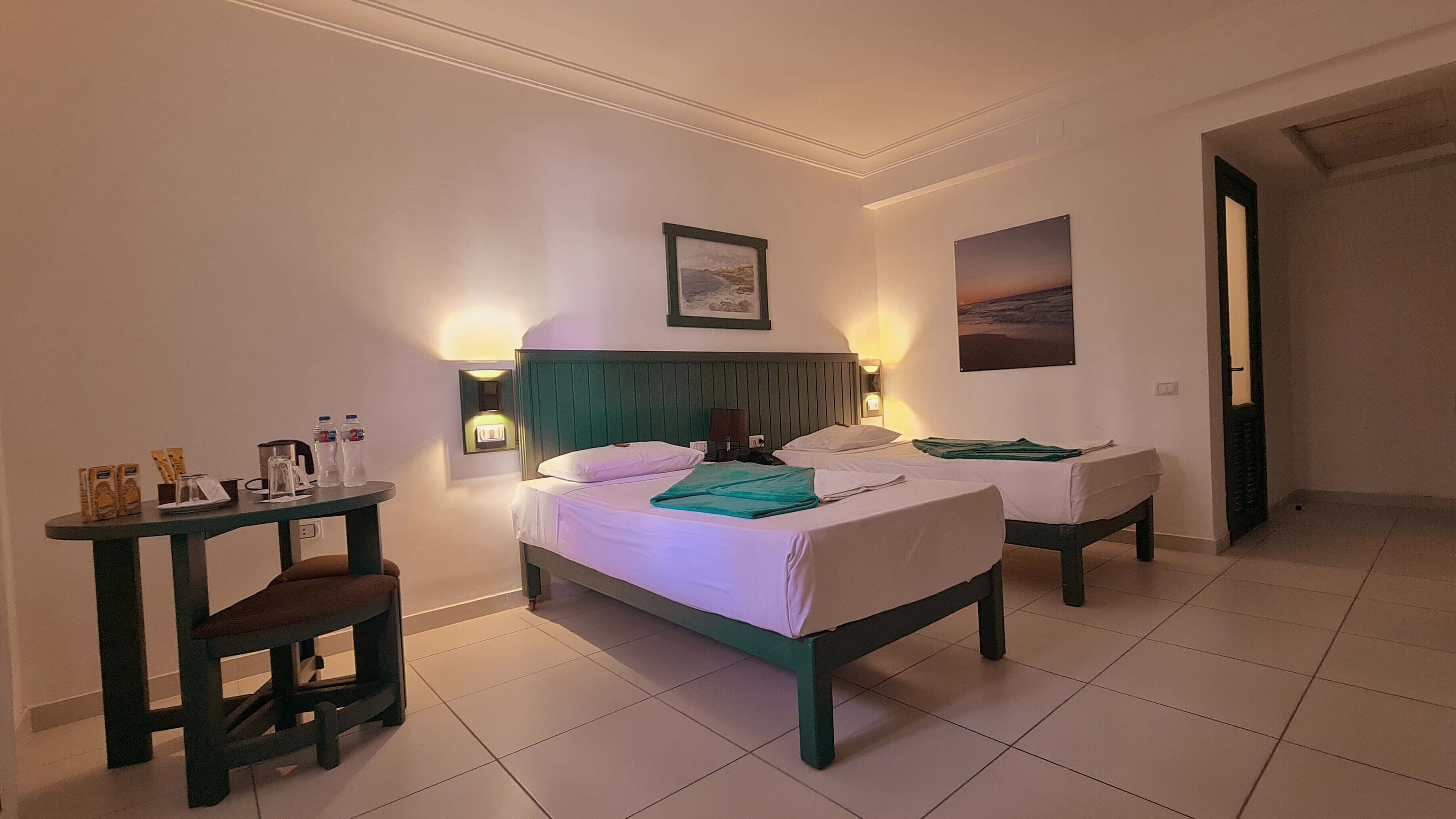 A hotel room with two beds, a small table with toiletries, and a view of the sea through the window.