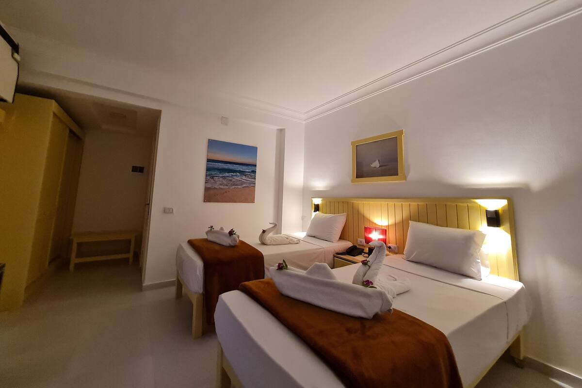 A hotel room with two single beds, a wooden headboard, a nightstand, a lamp, and a painting of a beach scene.