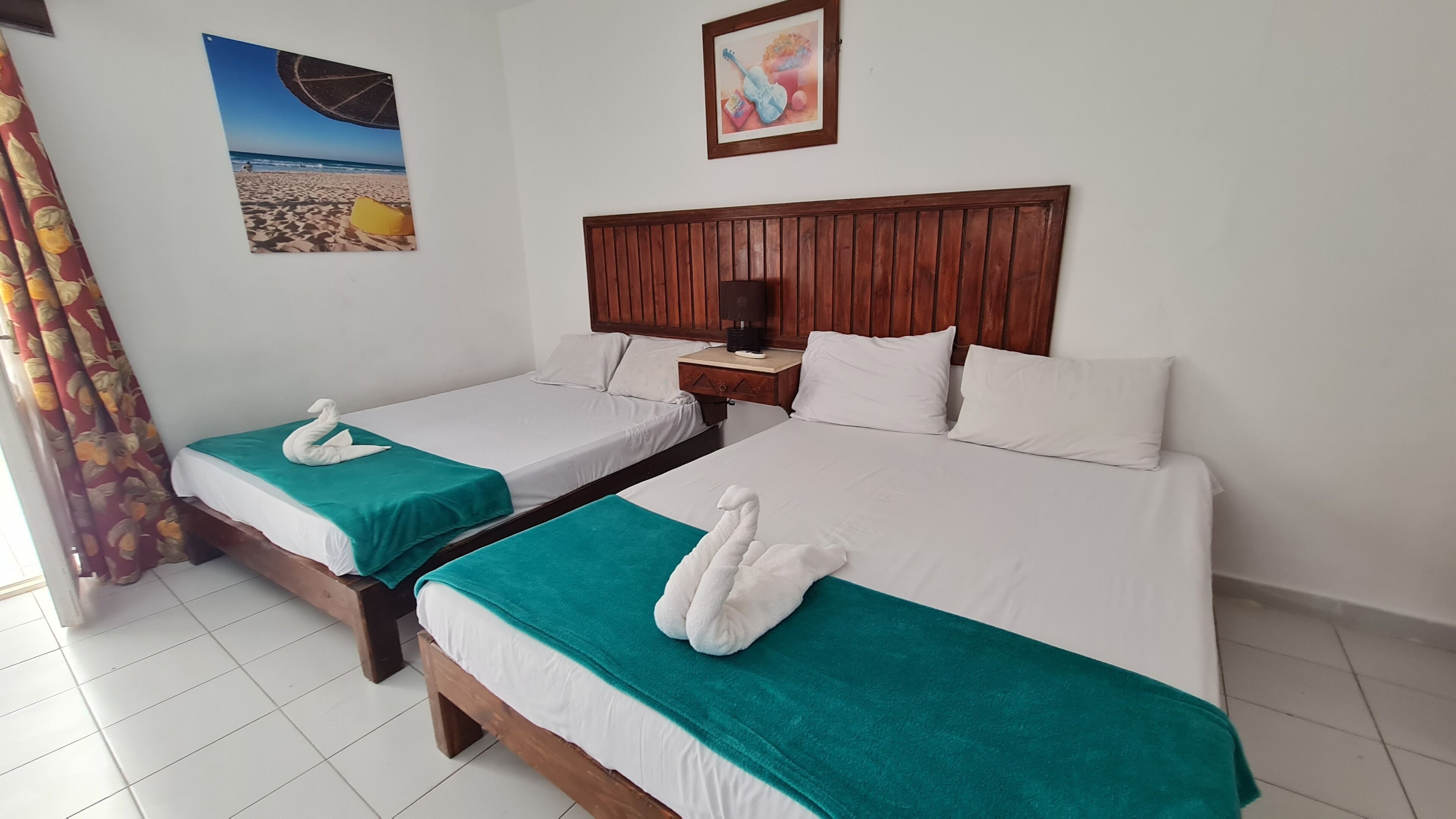 Habitación de hotel con dos camas, cada una con colcha color turquesa y almohadas blancas, y cabeceros de madera. En la pared, hay un cuadro con una escena de playa.