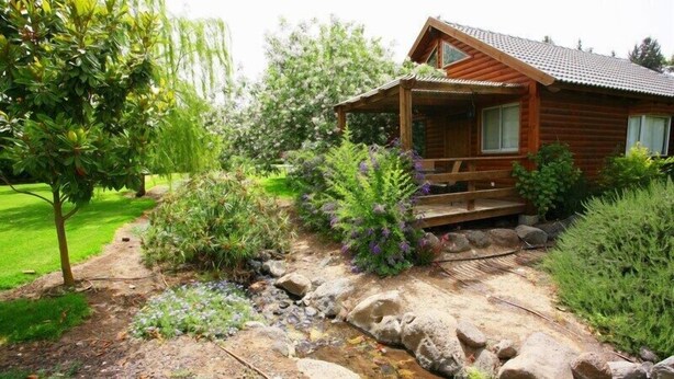A wooden cabin with a porch, surrounded by a garden with various plants and a small stream. - Nofey Habashan (HaGalil HaElyon)
