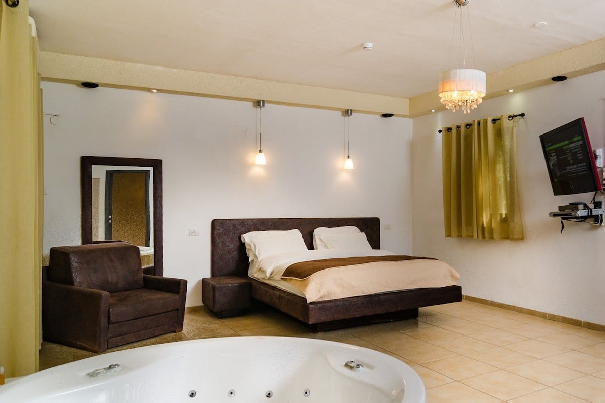 A bedroom with a bed, a chair, a large bathtub, and a chandelier.