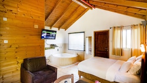 A wooden cabin room with a bed, a chair, a small table, a TV, a window with blinds, and a wooden door. - Nofey Habashan (HaGalil HaElyon)
