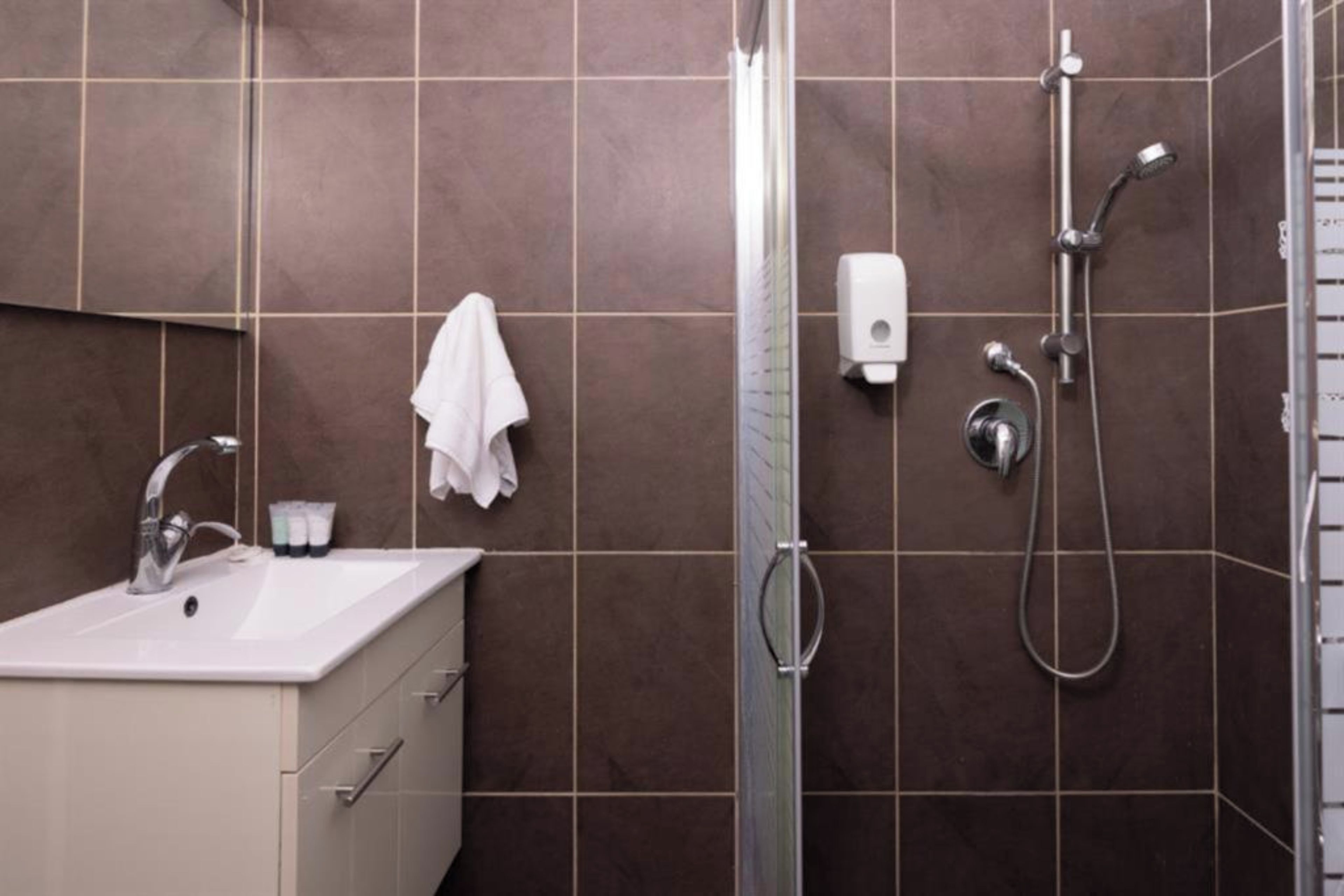 A bathroom with a sink, mirror, towel rack, shower, and a soap dispenser.
