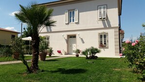 A two-story building with a white facade, a central entrance with a door, and two windows with shutters. There are potted plants and a lawn in front. - Villa Armonia (Lucca)