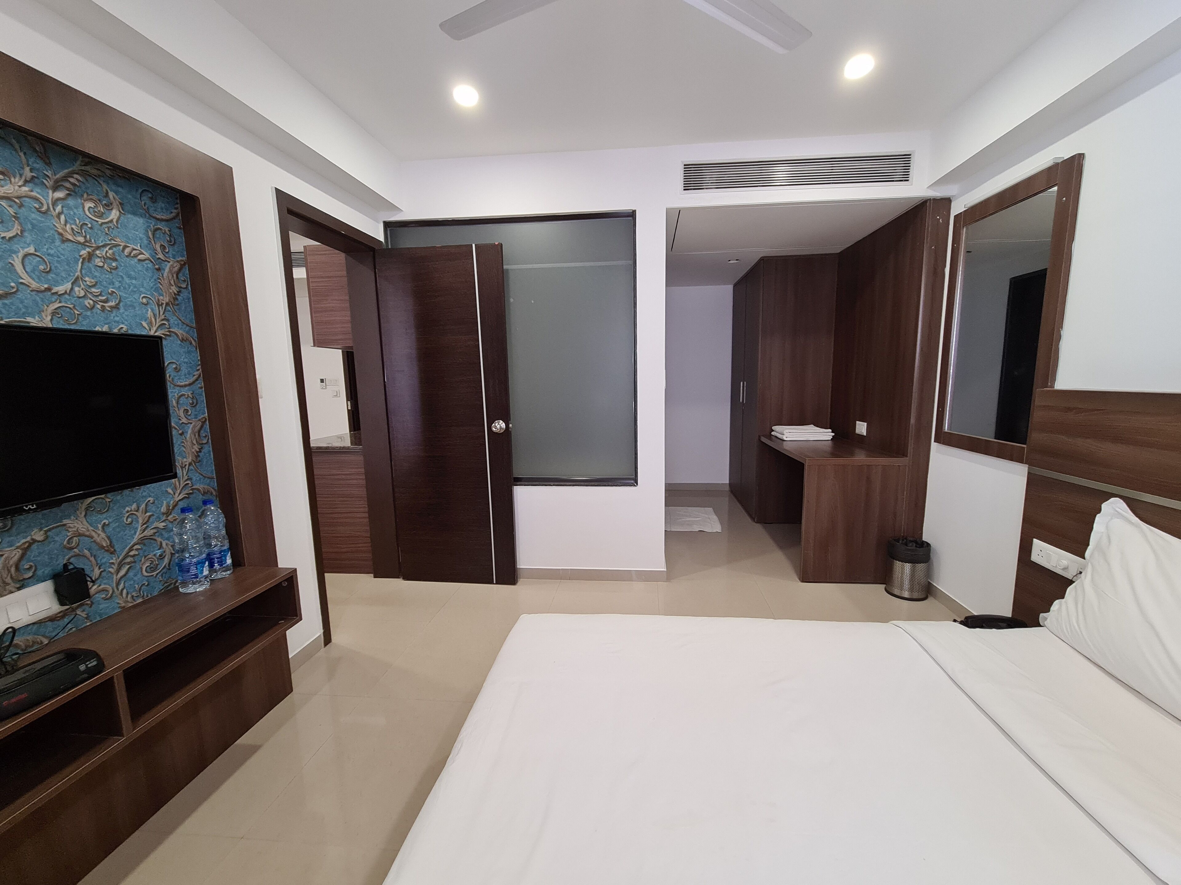 A modern hotel room with a large bed, a TV on a wooden stand, a bathroom with a sink and mirror, and a wardrobe with a sliding door.