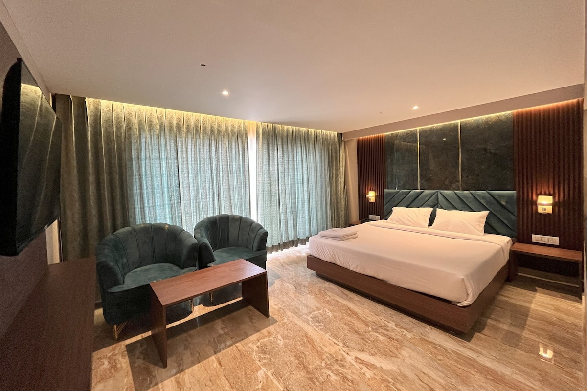 A modern hotel room with a large bed, two armchairs, a small table, and a large window with curtains.