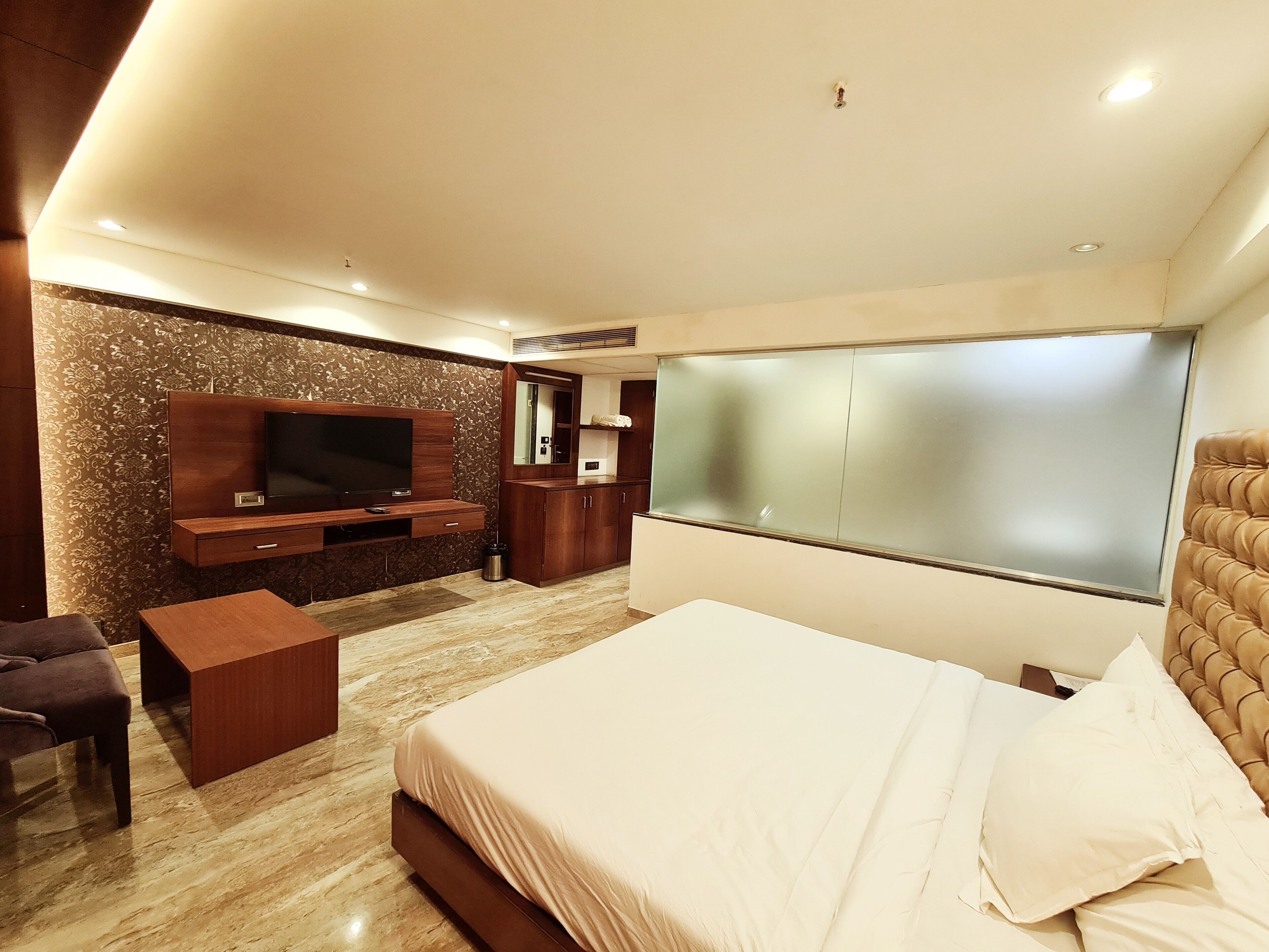 A modern bedroom with a large bed, a TV mounted on the wall, a small wooden side table, and a large window with frosted glass.