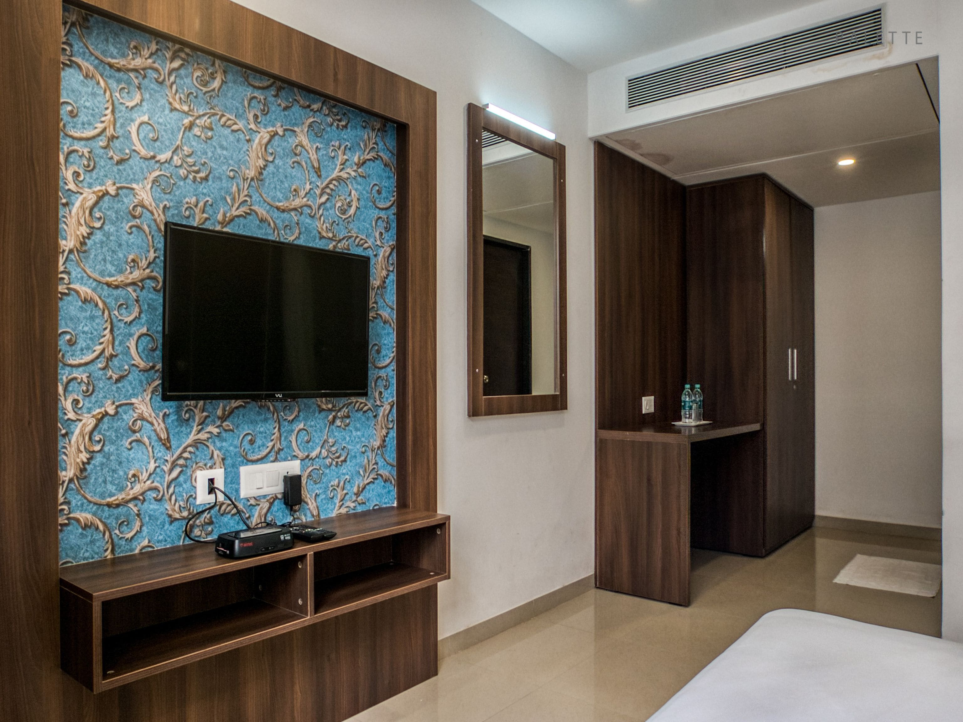 Ein modernes Hotelzimmer mit einem großen Flachbildfernseher an einer blauen Wand, einem hölzernen Schminktisch mit Spiegel und einem ordentlich bezogenen Bett.