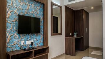 Ein modernes Hotelzimmer mit einem großen Flachbildfernseher an einer blauen Wand, einem hölzernen Schminktisch mit Spiegel und einem ordentlich bezogenen Bett.