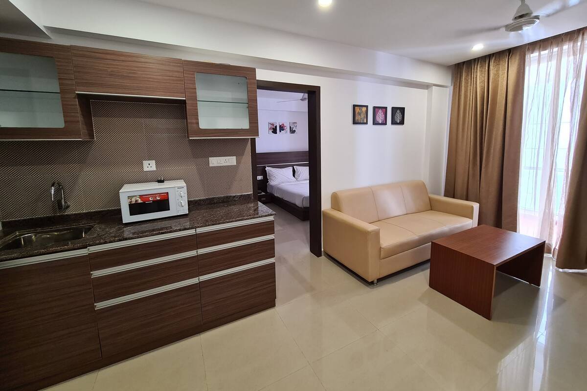 A modern kitchen with a microwave, a sink, and wooden cabinets. There is a beige sofa and a small wooden table in the living area.