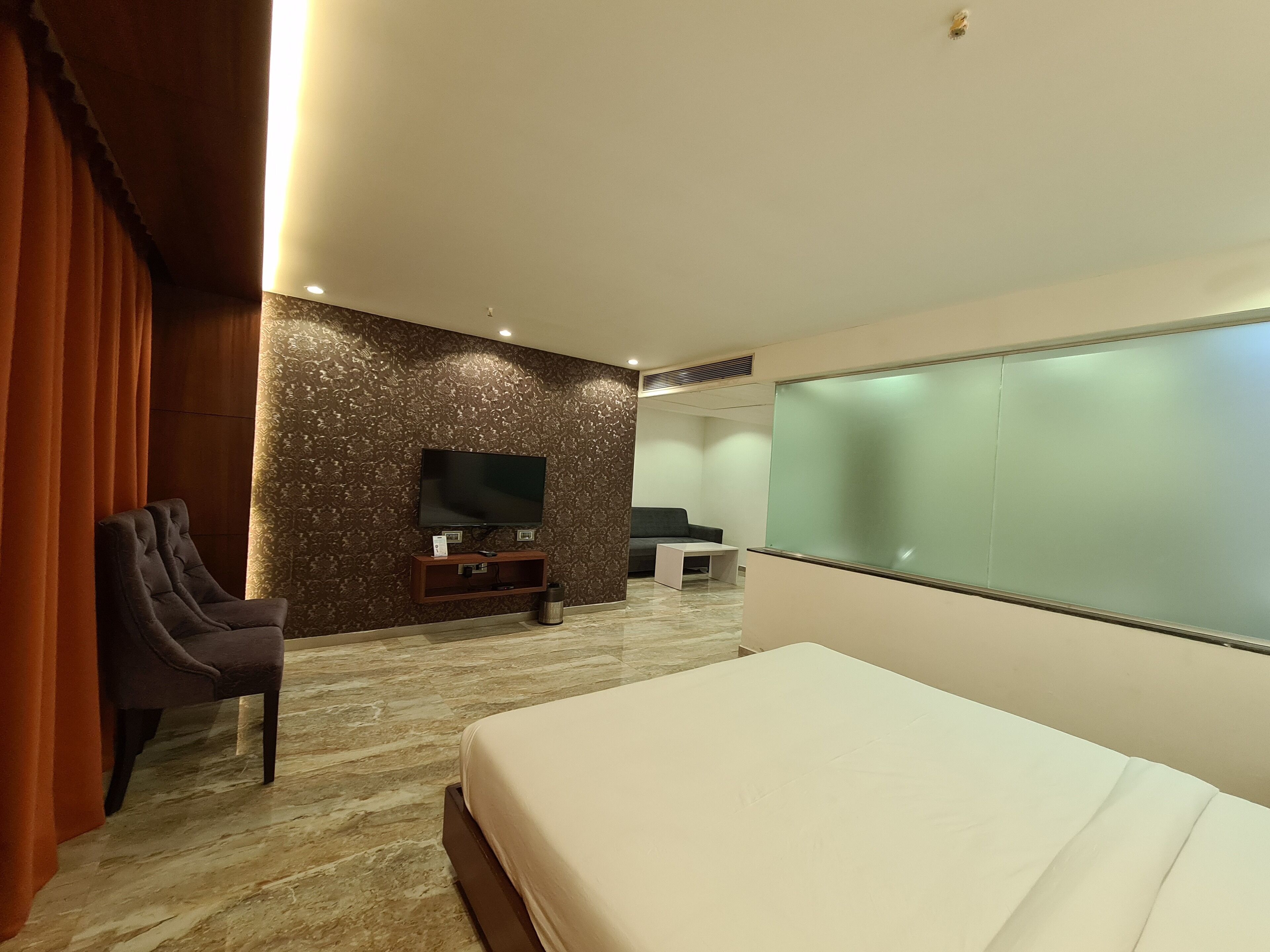 Una habitación de hotel moderna con una cama grande, un televisor de pantalla plana montado en una pared texturizada, una zona de estar con sofá y sillones, y una mesita.
