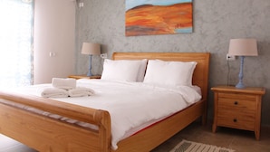 A neatly made bed with white linens and towels, a wooden headboard, two bedside lamps, a small wooden nightstand, and a painting on the wall. - Nahal Faran 32 (Ramat Negev)