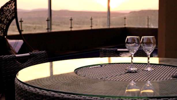 A round table with two wine glasses on a woven tablecloth, overlooking a scenic view with mountains and a sunset. - Nahal Faran 32 (Ramat Negev)