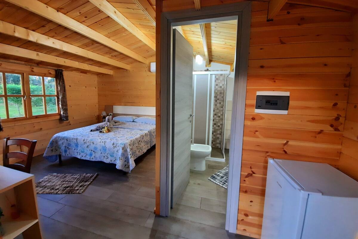 A wooden cabin with a bed, a small table, and a chair. There is a bathroom visible through a doorway.