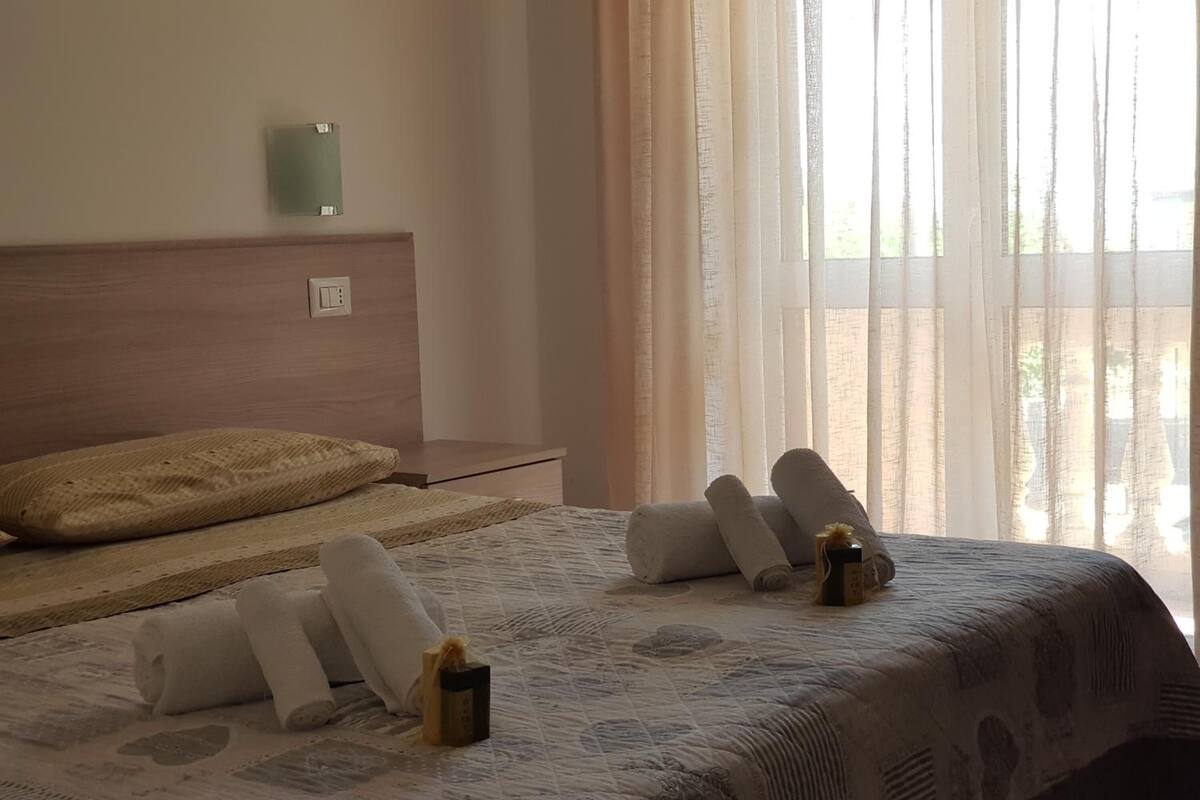 A hotel room with a bed, a wooden headboard, a bedside table, and a window with sheer curtains.