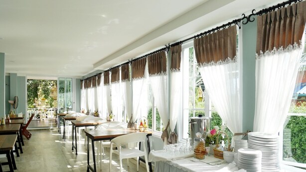 A long dining room with tables and chairs, large windows with curtains, and a view of greenery outside. - Sky Cottage (Rayong)