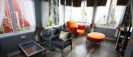 A room with a ceiling fan, a glass coffee table, two sofas, an orange ottoman, and large windows with curtains.