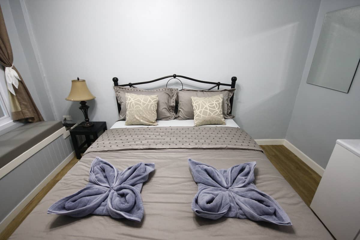A neatly made bed with two towels folded into butterfly shapes, a lamp, and a window with curtains.