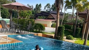 A swimming pool with a person swimming, surrounded by palm trees and lounge chairs. - Santorini Khanom (Khanom)