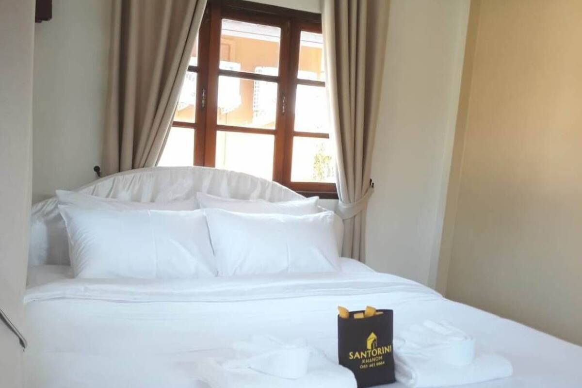 A neatly made bed with white linens, a black Santorini sign, and beige curtains.
