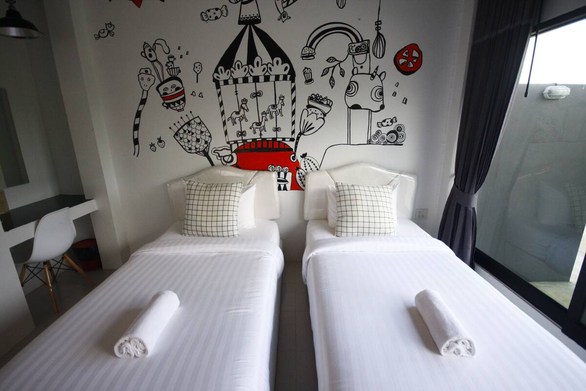 Habitación de hotel con dos camas, ropa de cama blanca y un mural con diversas ilustraciones en blanco y negro en la pared.