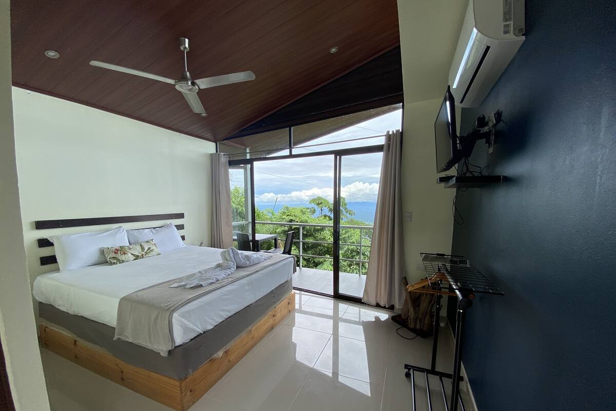 A bedroom with a bed, a ceiling fan, a TV, and a balcony with a view of the ocean.