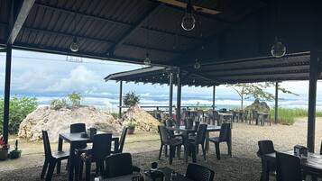 An outdoor dining area with tables and chairs, a covered roof, and a view of the ocean.