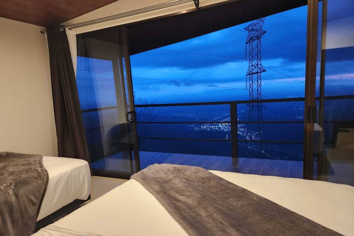 A hotel room with two beds, a large window offering a view of a power line tower and distant landscape.