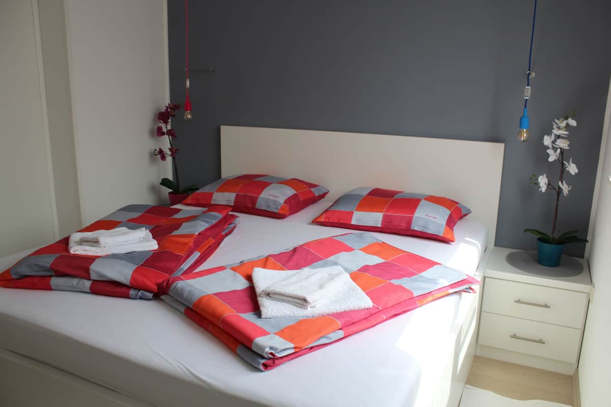 A neatly made bed with a checkered bedspread, two pillows, and folded towels. There is a bedside table with a plant and a lamp.