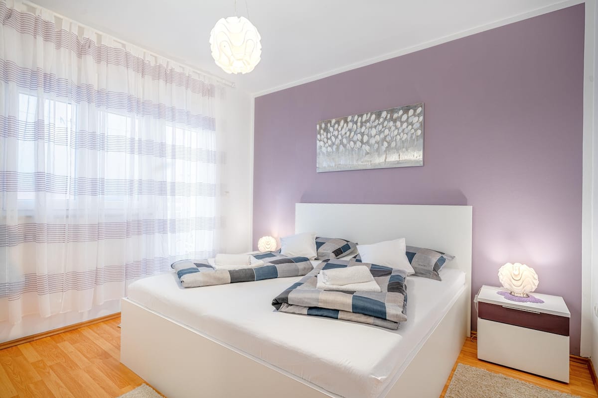 A bedroom with a bed, bedside tables, a nightstand, a wall-mounted light fixture, and a window with sheer curtains.