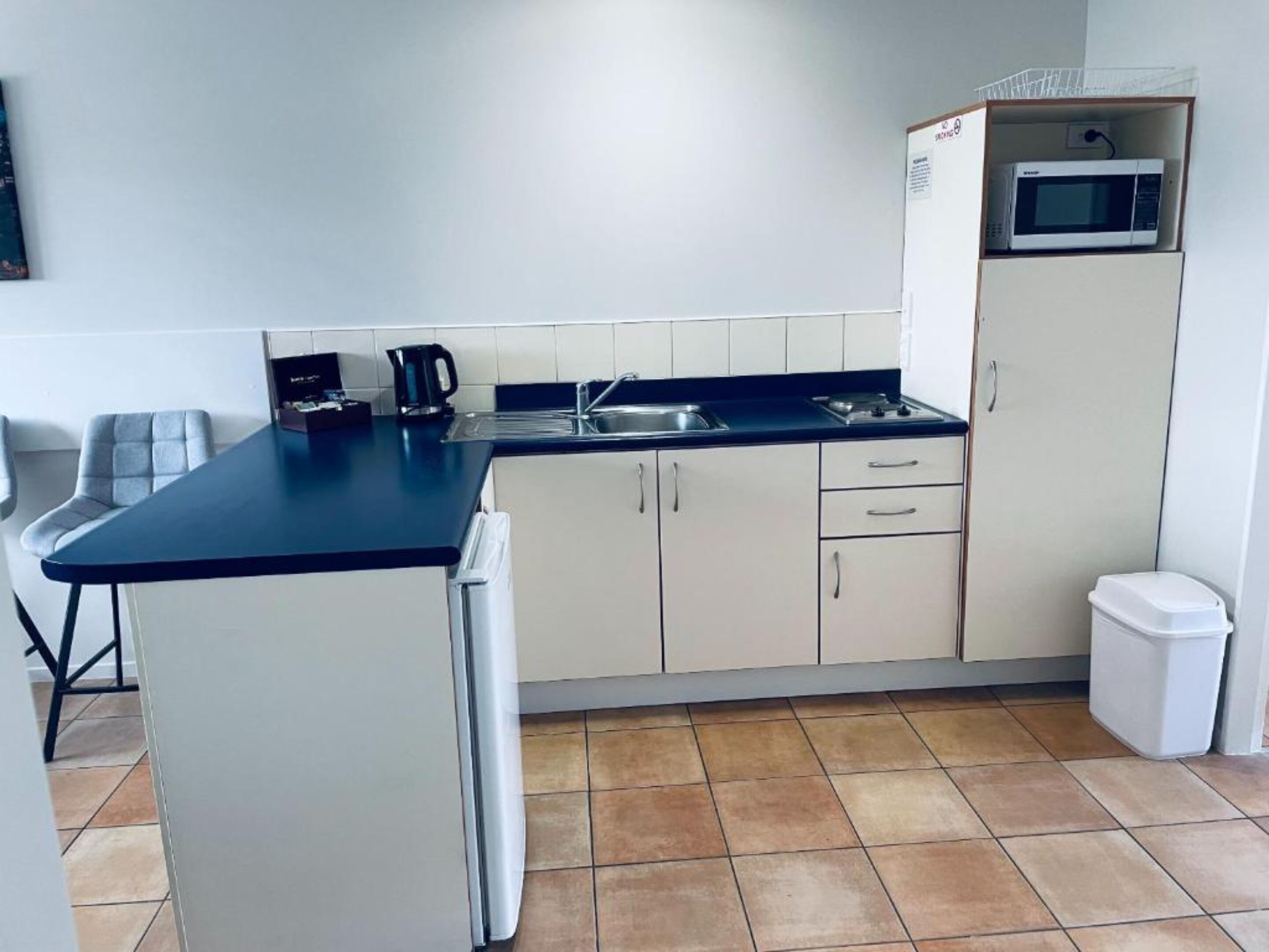Una cocina compacta con refrigerador, microondas, lavamanos y un pequeño comedor con una silla.