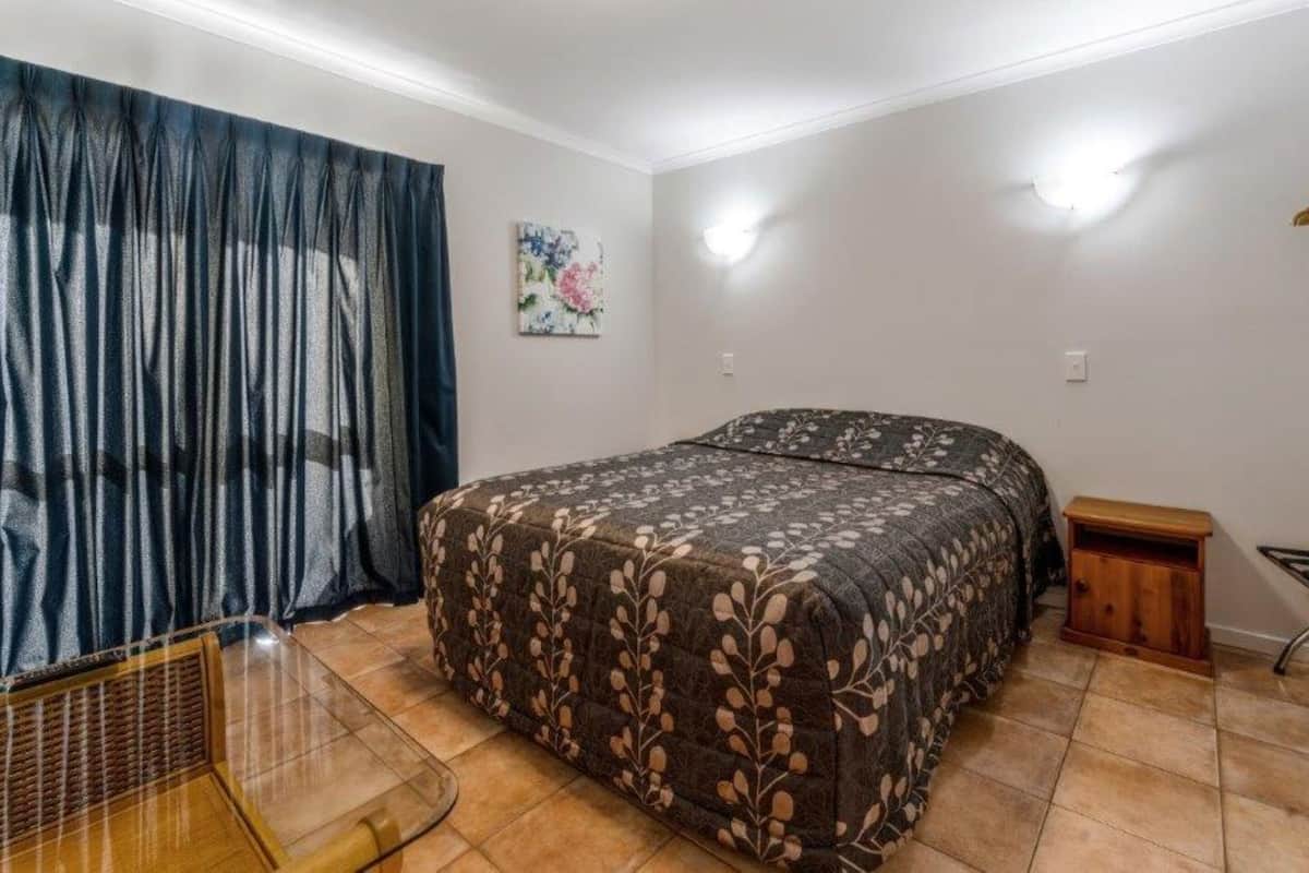 A bedroom with a bed, a small wooden nightstand, a chair, and a window with sheer curtains.