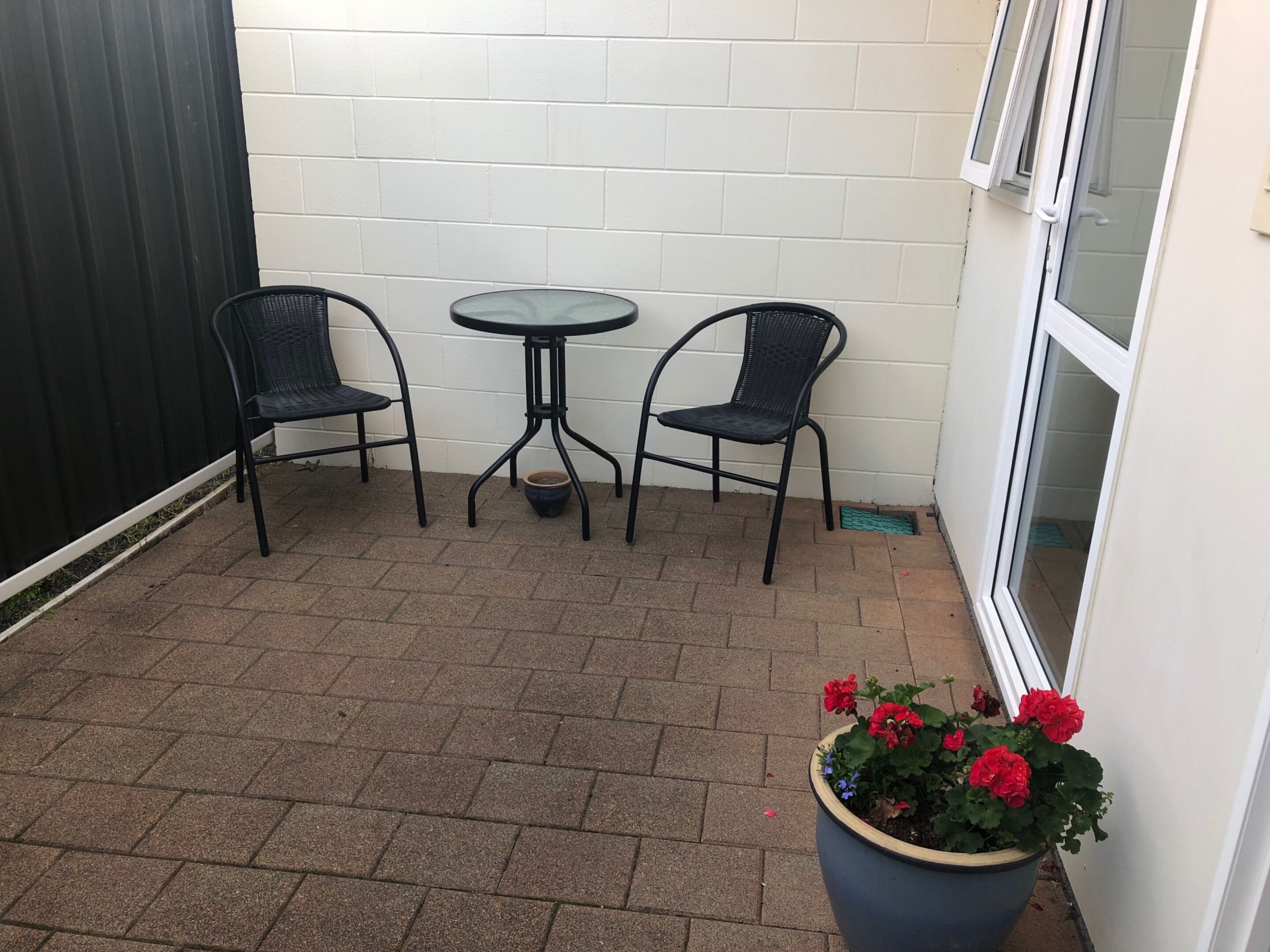 A small patio area with two black metal chairs and a round table, a potted plant with red flowers, and a white brick wall.
