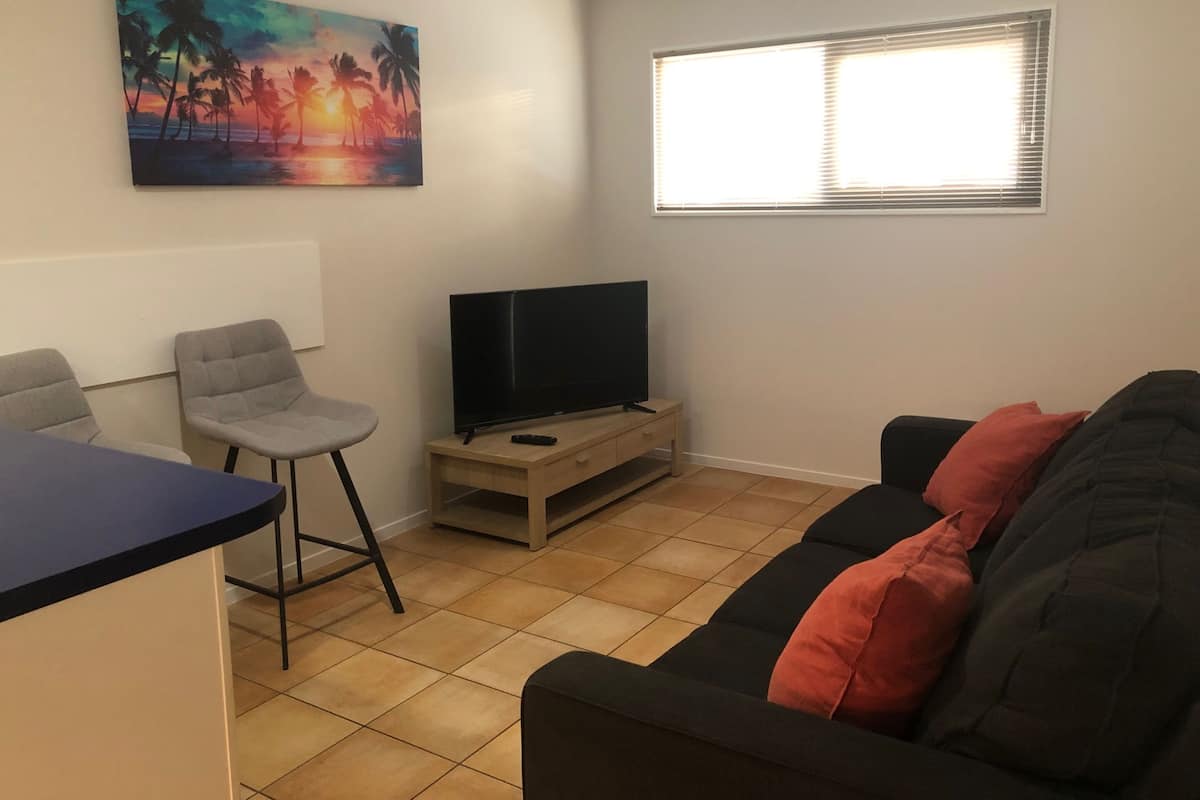 A living room with a flat-screen TV on a stand, a couch with red cushions, a painting of a sunset, and a window with blinds.