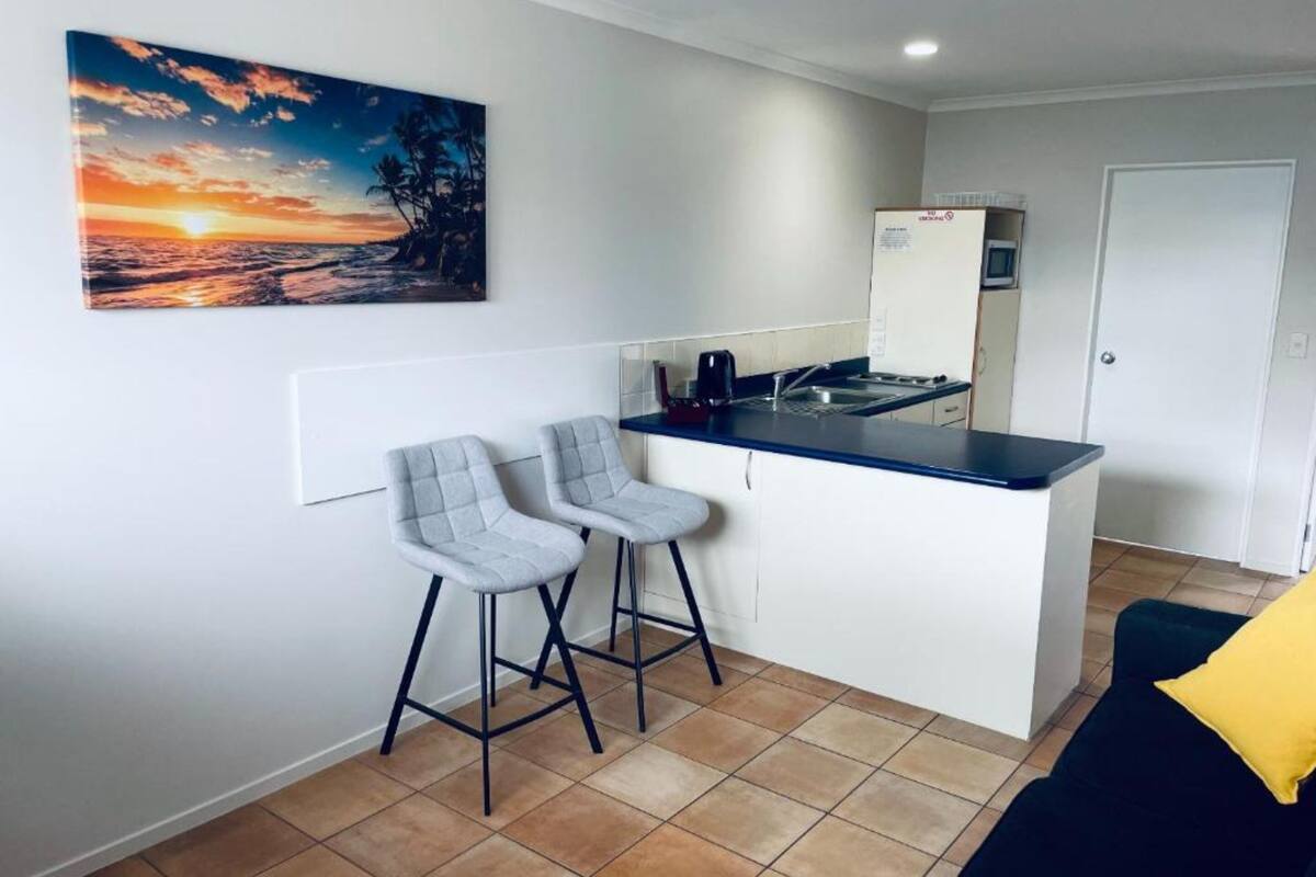 A compact kitchen with two bar stools, a small island, and a painting of a sunset.