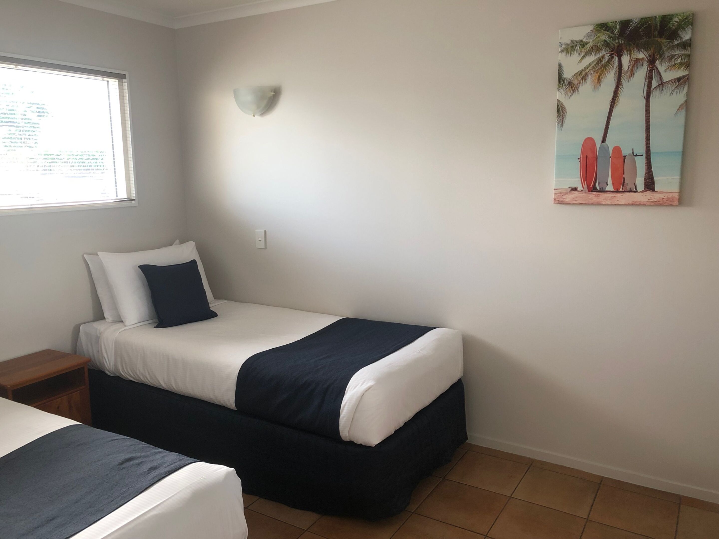 A single bed with a dark blue and white bedspread, a nightstand, a wall-mounted light, and a beach-themed painting.