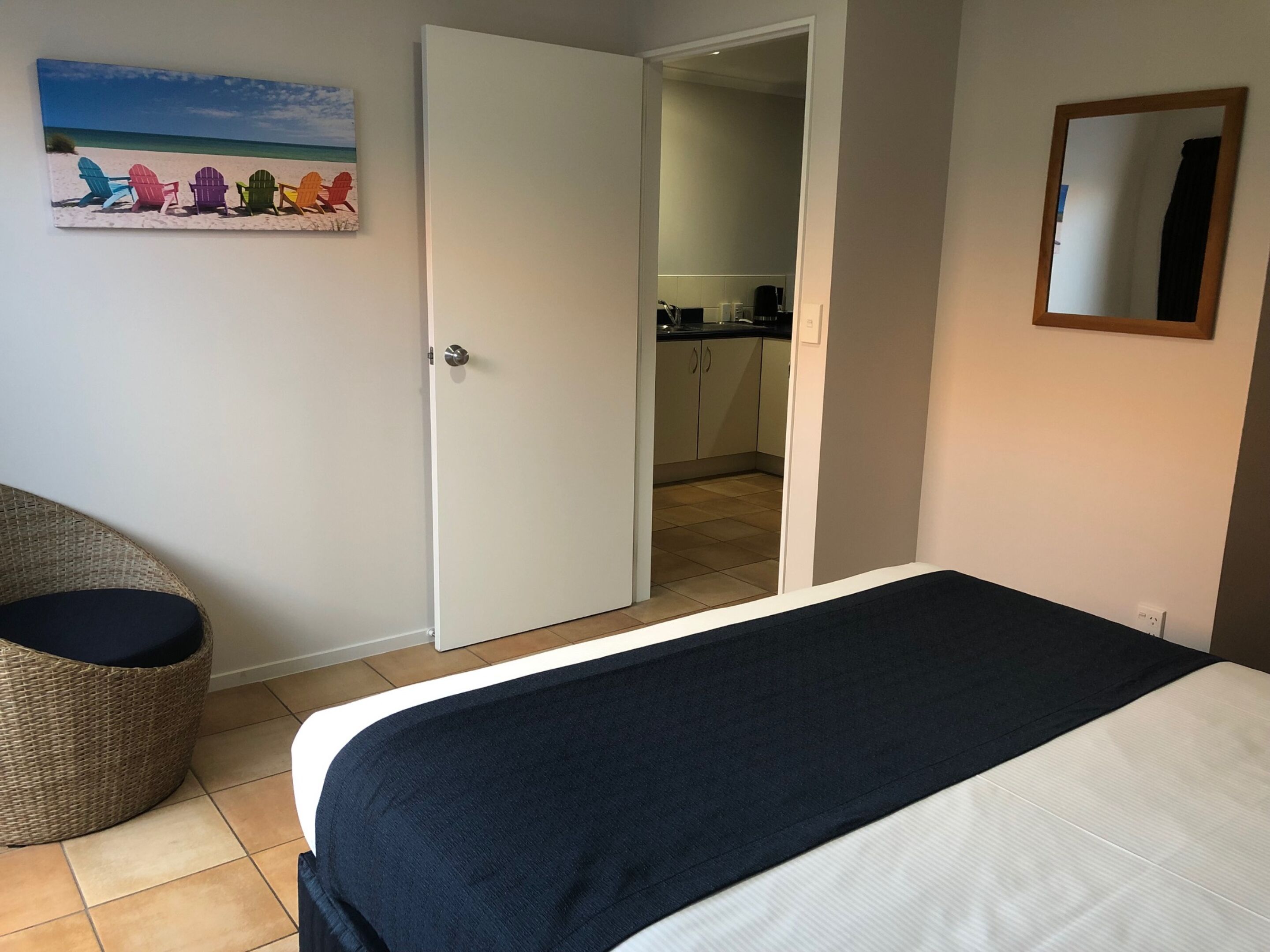 Une chambre avec un lit, une chaise en osier, une porte donnant sur une cuisine et un tableau sur le thème de la plage accroché au mur.