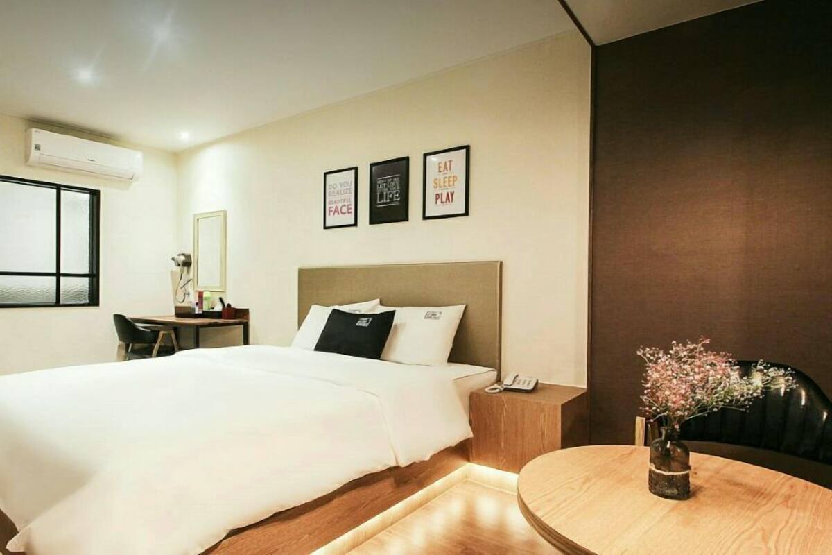 Habitación de hotel con cama, mesita de noche, escritorio, silla y una pequeña mesa redonda con un florero.