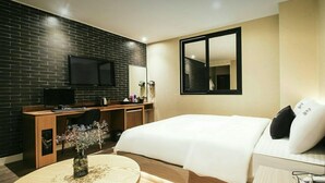 A modern hotel room with a brick wall, a flat-screen TV, a desk with a chair, a bed with white bedding, and a large mirror. - The Hotel Changwon (Changwon)