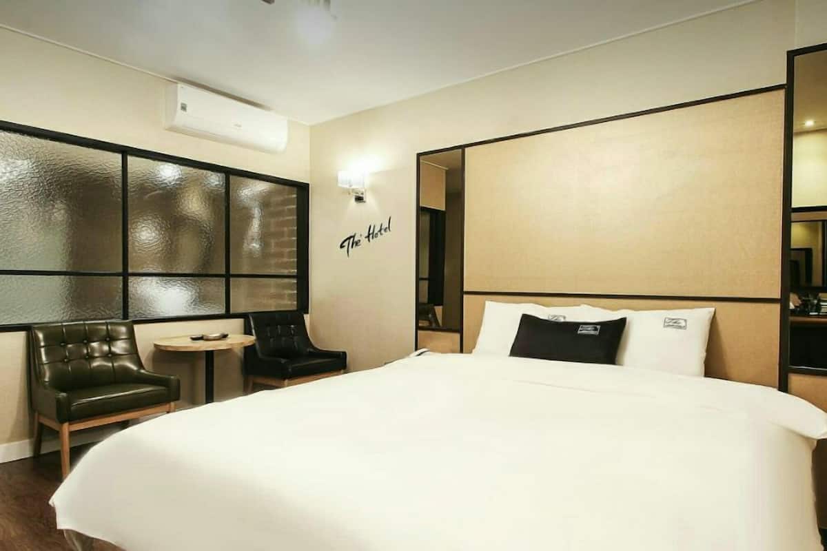 Habitación de hotel con una cama grande, un rincón de descanso con una silla y una mesita, aire acondicionado en la pared y un espejo.