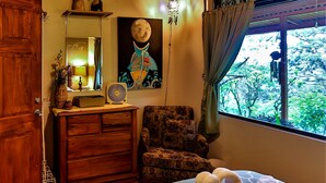 Un dormitorio con techo de madera, una cama con colcha verde y blanca estampada, un aparador de madera, una silla, una lámpara de pared, un cuadro y una ventana con cortinas.