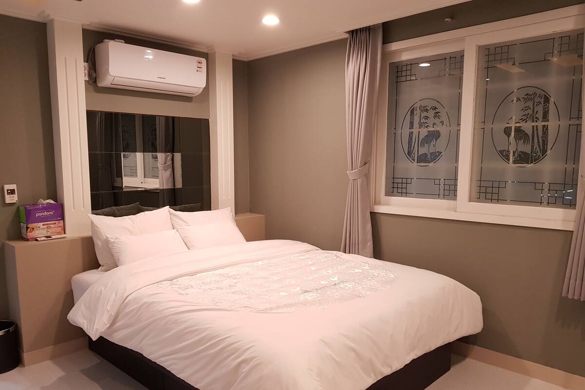 Ein ordentlich bezogenes Bett mit weißen Leinen, eine Wand-Klimaanlage, ein Fenster mit dekorierter Scheibe und ein kleiner Nachttisch mit Taschentuchbox.