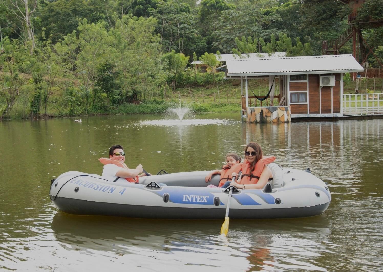A family on an inflatable raft with "INTEX" branding, floating on a lake with a dock and a building in the background.