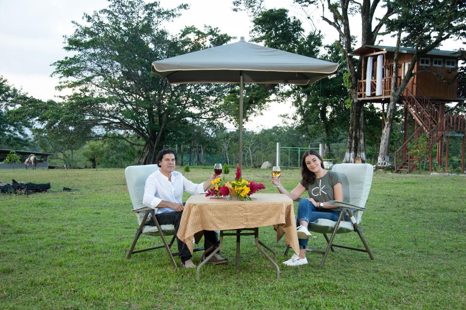 Dos personas sentadas en una mesa al aire libre, bajo una toldilla, rodeadas de follaje y una estructura de madera al fondo.