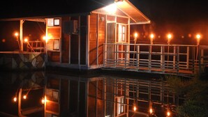 A wooden cabin with a balcony, a small porch, and a reflection in the water.