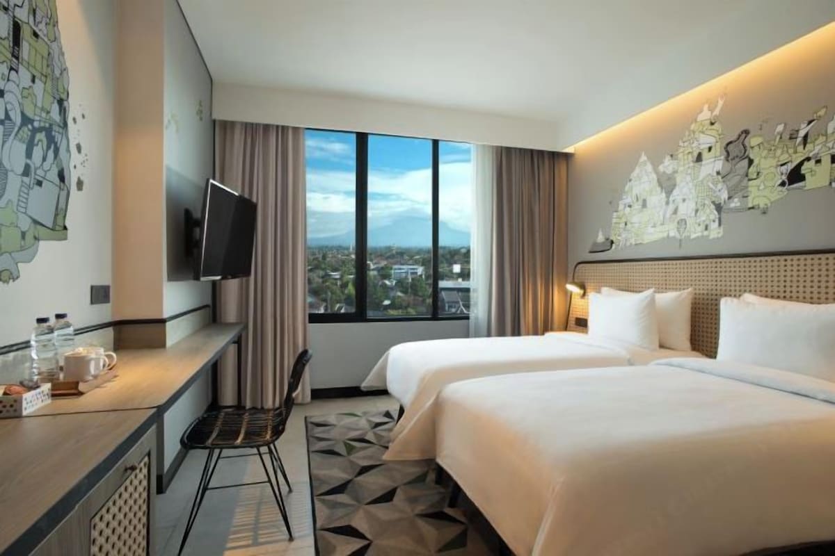 A hotel room with two beds, a TV on the wall, a desk with a chair, and a large window offering a city view.