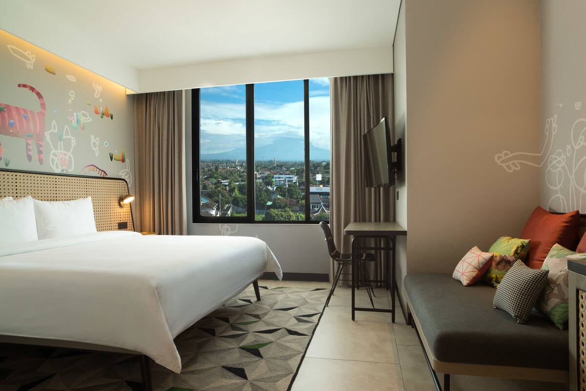A hotel room with a large bed, a sofa, a desk, and a TV. There is a window with a city view.