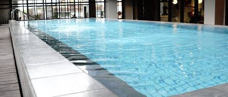 An indoor swimming pool with clear water, surrounded by a tiled deck and glass walls.