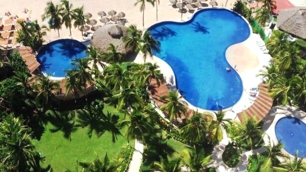 Eine Vogelperspektive zeigt ein Resort mit zwei großen Pools, umgeben von Palmen und einem Sandstrand.